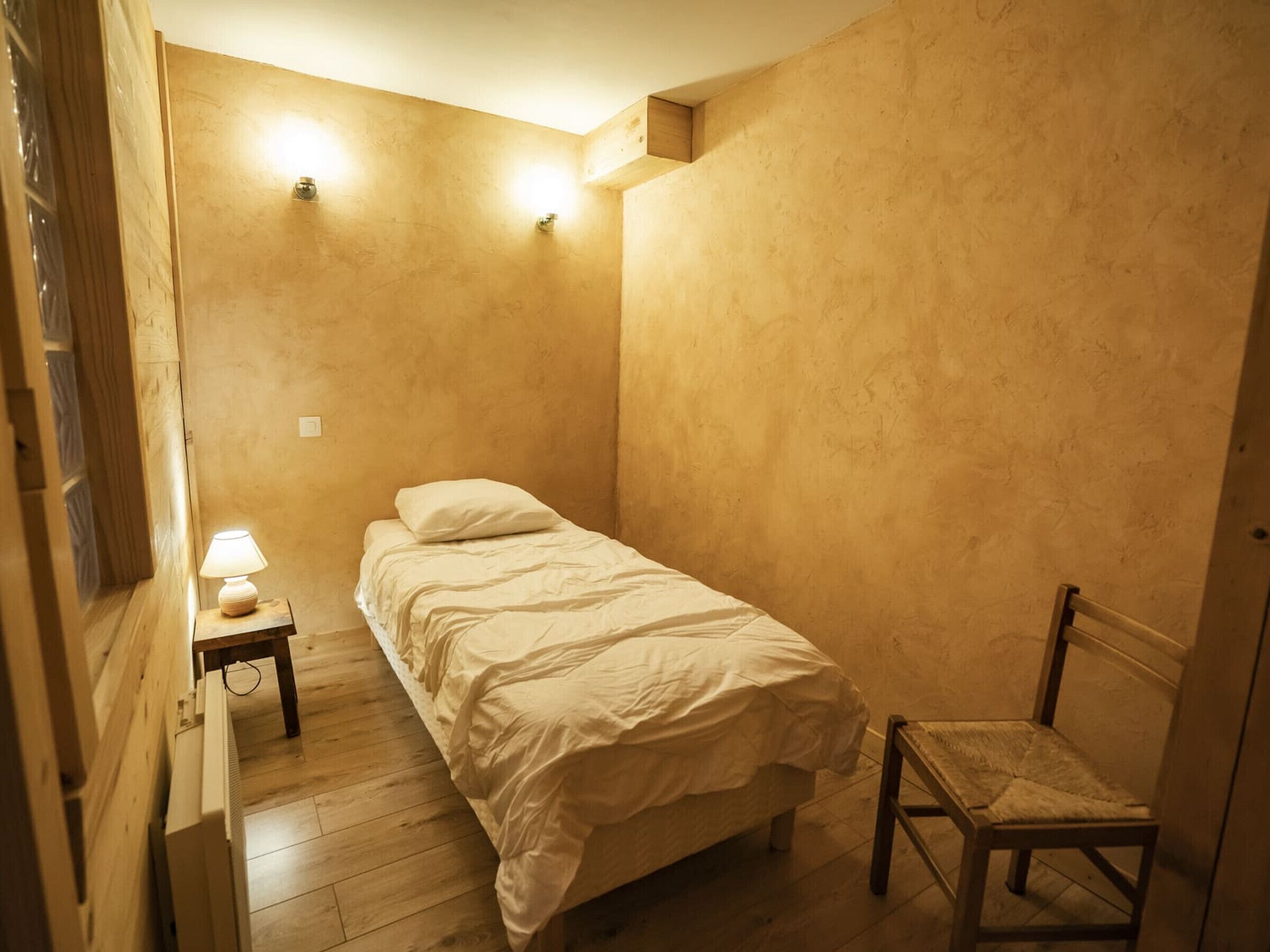 A small, cozy bedroom with a single bed, a nightstand with a lamp, and a chair.