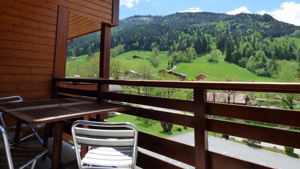A wooden balcony with a table and chair, overlooking a green valley and forested hills. - Charming 3-room south with balcony near cable cars, 6 ppl (Le Grand-Bornand)