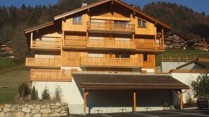 A multi-story wooden chalet with balconies, a covered parking area, and a stone wall in front. - Charming 3-room south with balcony near cable cars, 6 ppl (Le Grand-Bornand)