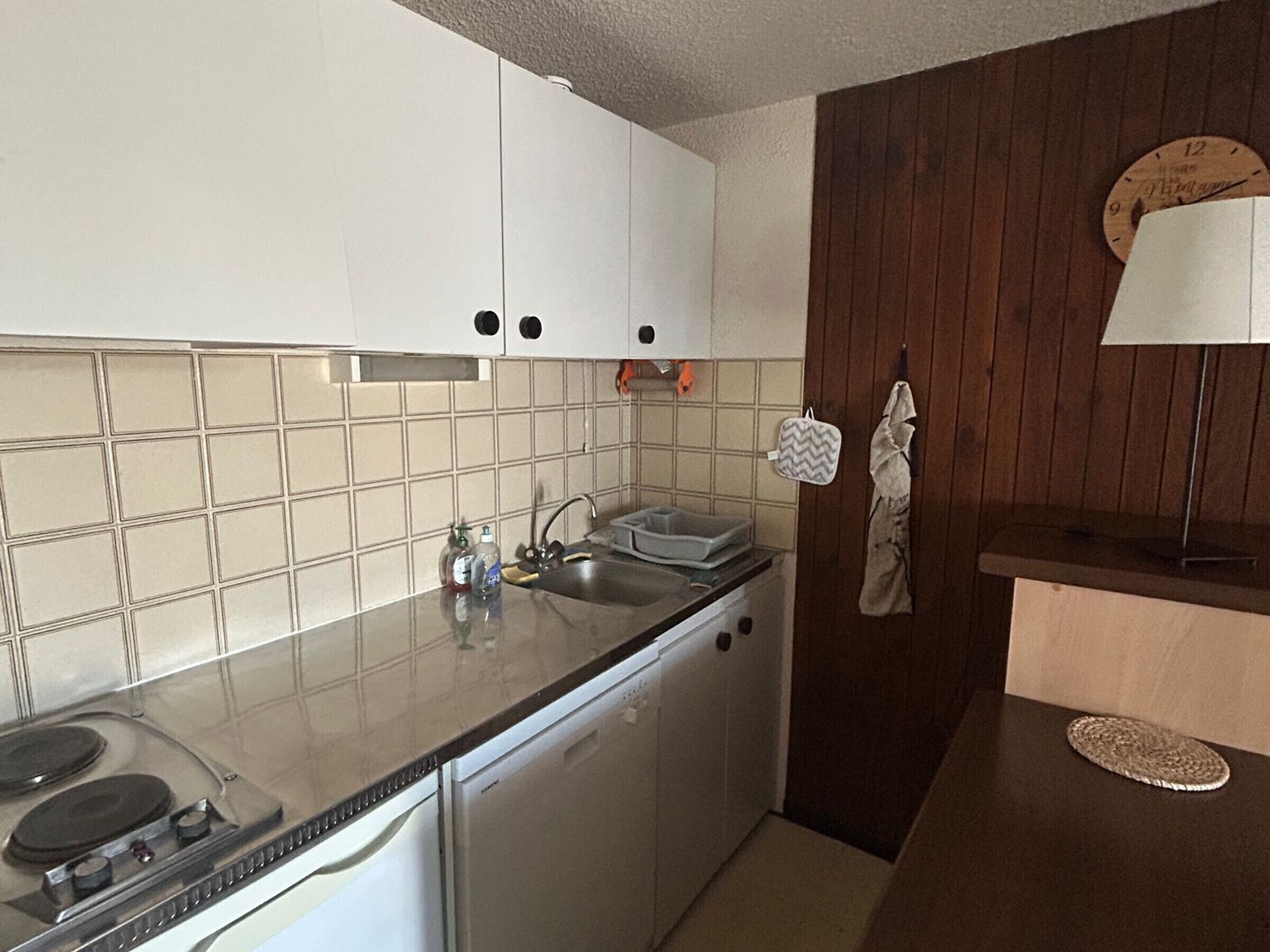 Una cocina compacta con armarios blancos, un lavamanos de acero inoxidable, lavavajillas y un revestimiento de pared de madera.