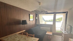 Ein Zimmer mit Holztisch, einer Couch, einem Fernseher und einem Fenster mit Blick auf Grünflächen.