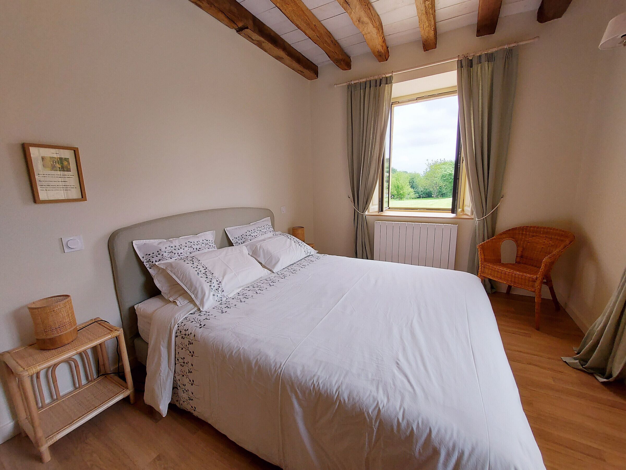 A bedroom with a bed, a wicker chair, a window with curtains, and a radiator.