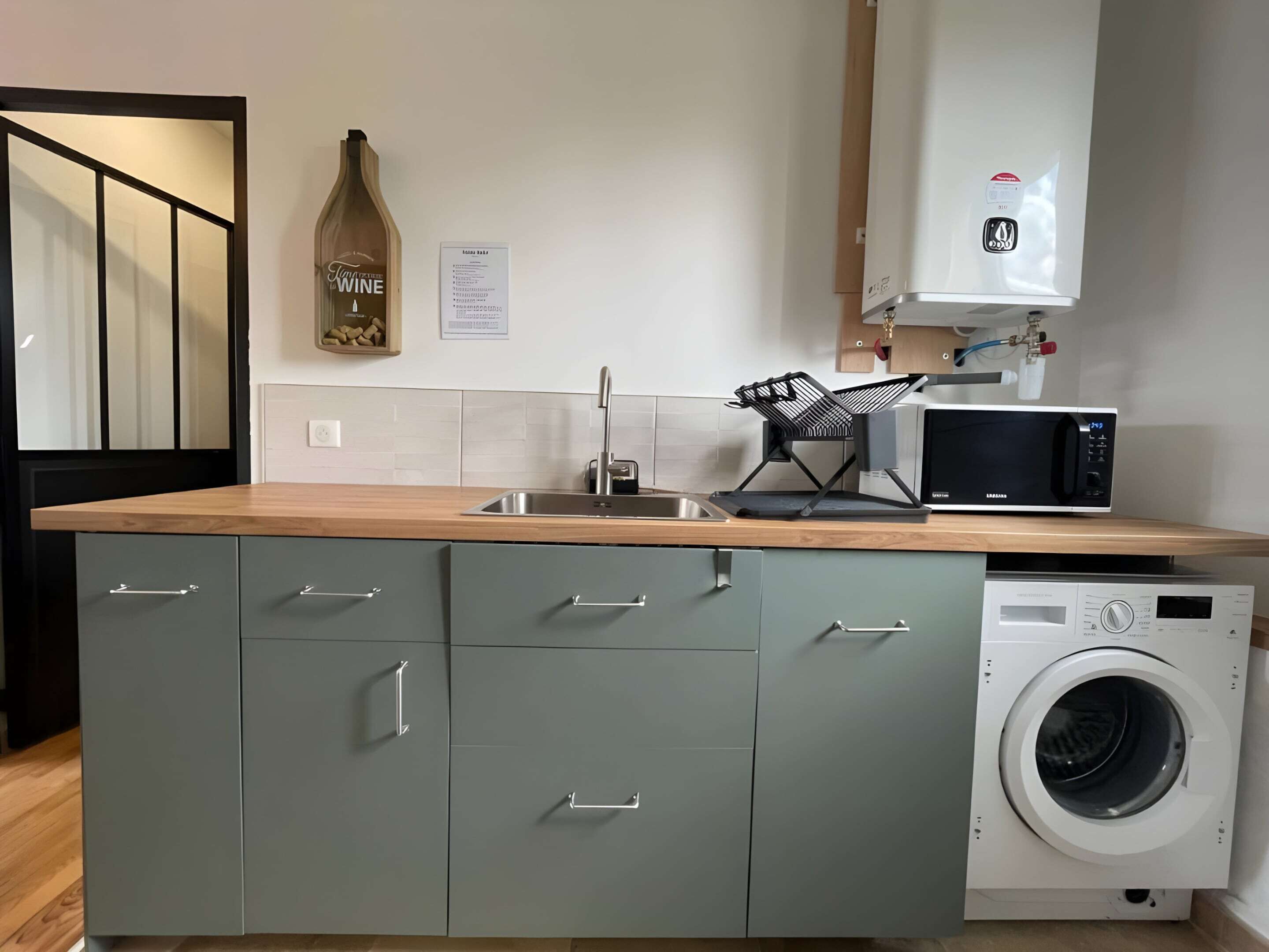 A compact kitchen with a built-in washing machine, a microwave, and a wine bottle on the counter.