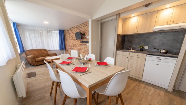 A modern kitchen with white cabinets, a built-in stove, and a dishwasher. There is a dining area with a wooden table and white chairs, and a living room with a brown sofa and a TV mounted on the wall. - Chalet in Normandy Near Veules-les-roses (Veules-les-Roses)