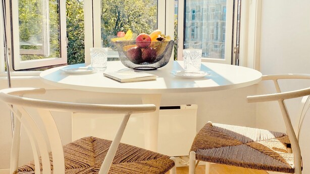 Un comedor con una mesa redonda, dos sillas de mimbre y un cuenco de fruta sobre la mesa.