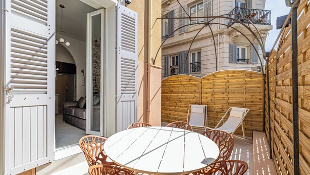 Un patio al aire libre con una mesa redonda y sillas de mimbre, una valla de madera y vistas a un edificio con balcones.