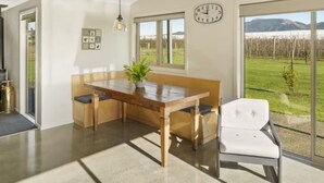A modern dining area with a wooden table, bench seating, and a white chair. A clock is visible on the wall, and there is a view of a grassy field and mountains through large windows. - Relax in Riwaka, Sun, Space, Cycle (Riwaka)