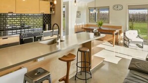A modern kitchen with a central island, bar stools, and a view of a garden through large windows. - Relax in Riwaka, Sun, Space, Cycle (Riwaka)