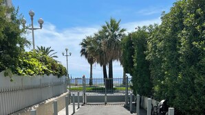 A paved pathway leading to a gated entrance with a view of the sea and palm trees.