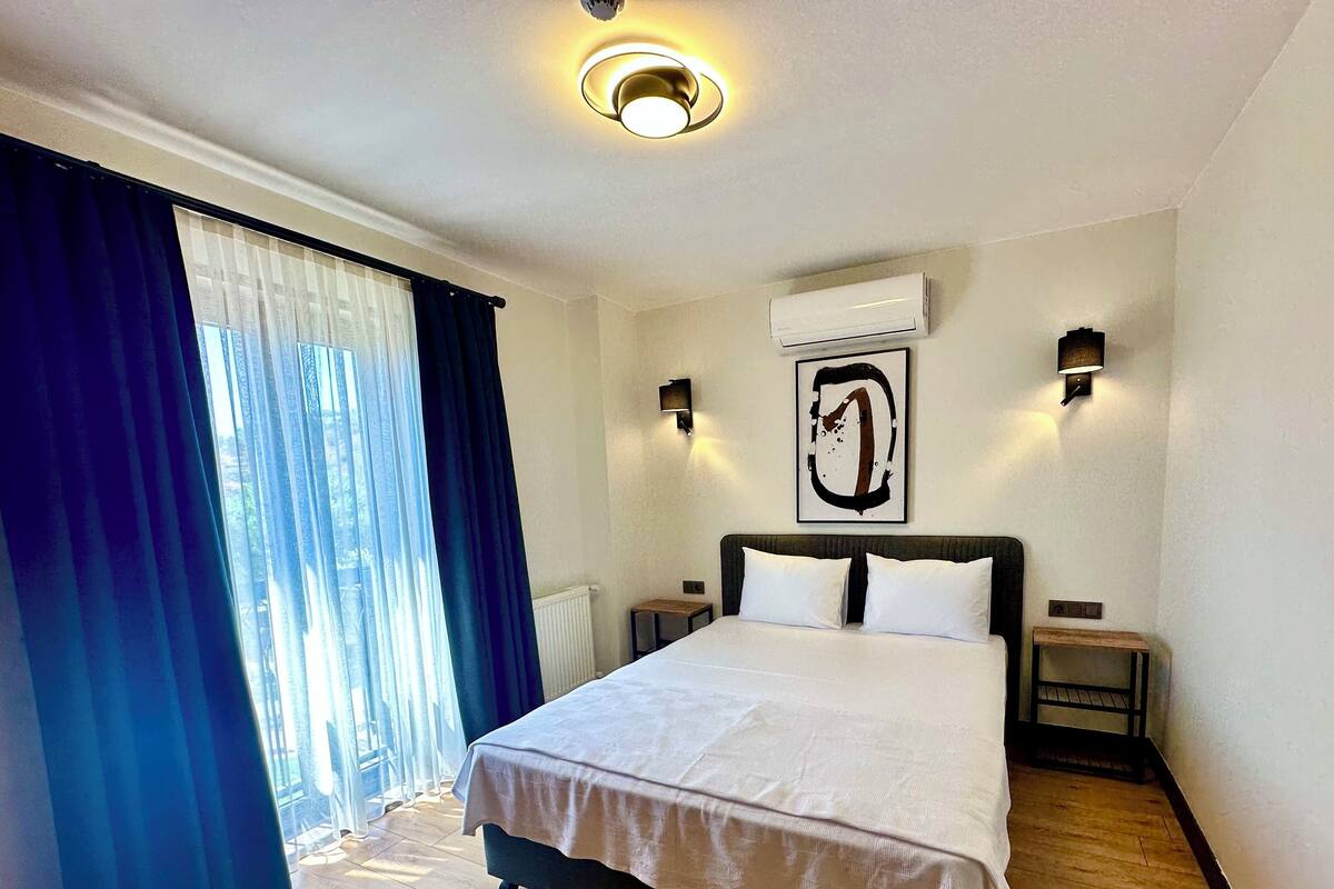 Habitación de hotel con una cama grande, dos mesitas de noche, un equipo de aire acondicionado y una ventana con cortinas azules.