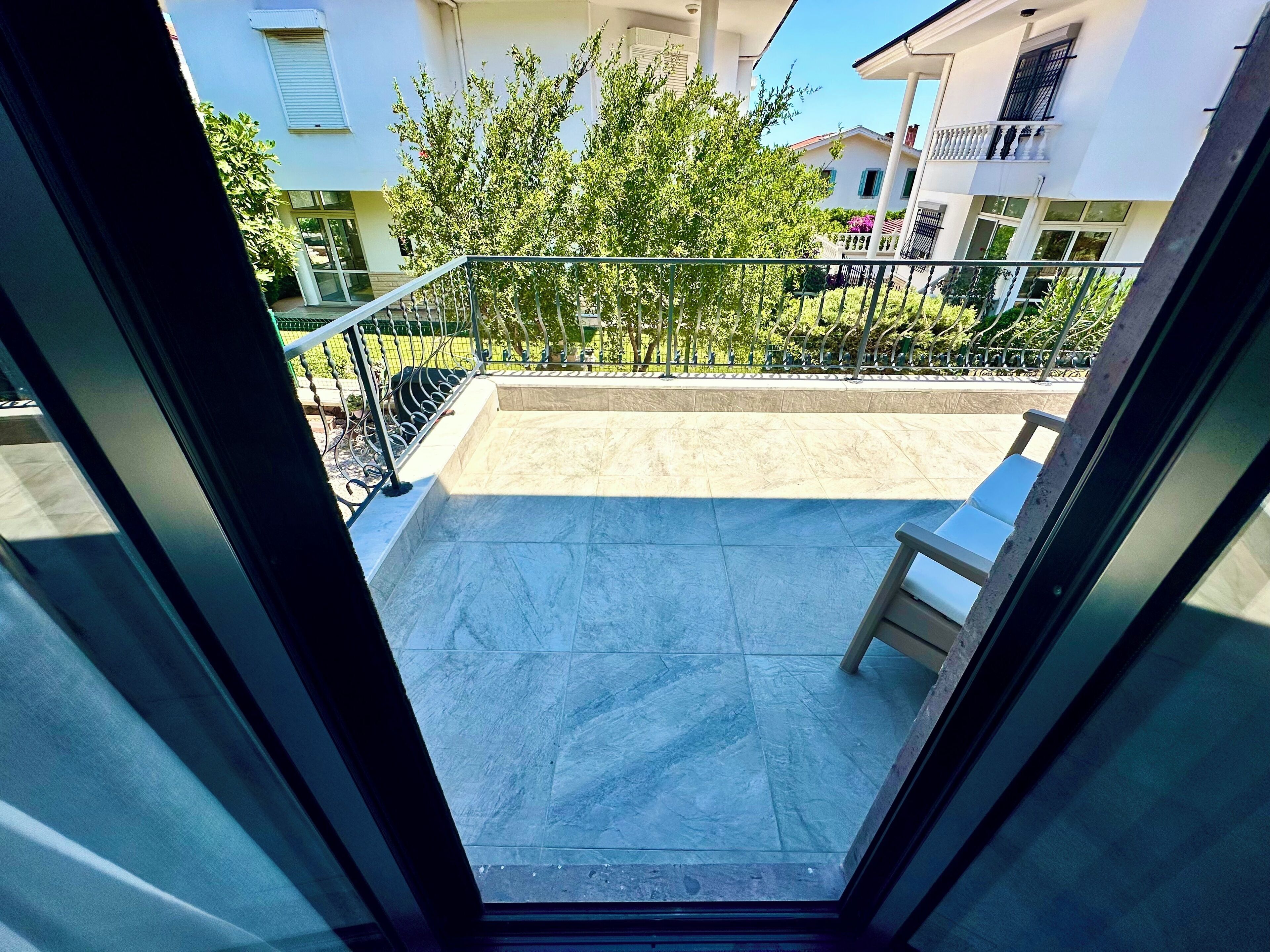 Un balcon doté d’une rambarde en métal, d’une chaise et offrant une vue sur les bâtiments voisins et la verdure.
