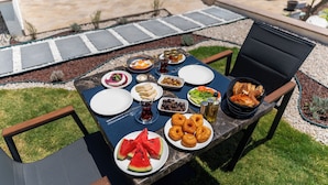 Une table à manger installée à l’extérieur, sur laquelle on trouve divers plats, notamment des tranches de pastèque, des beignets et un bol d’olives.