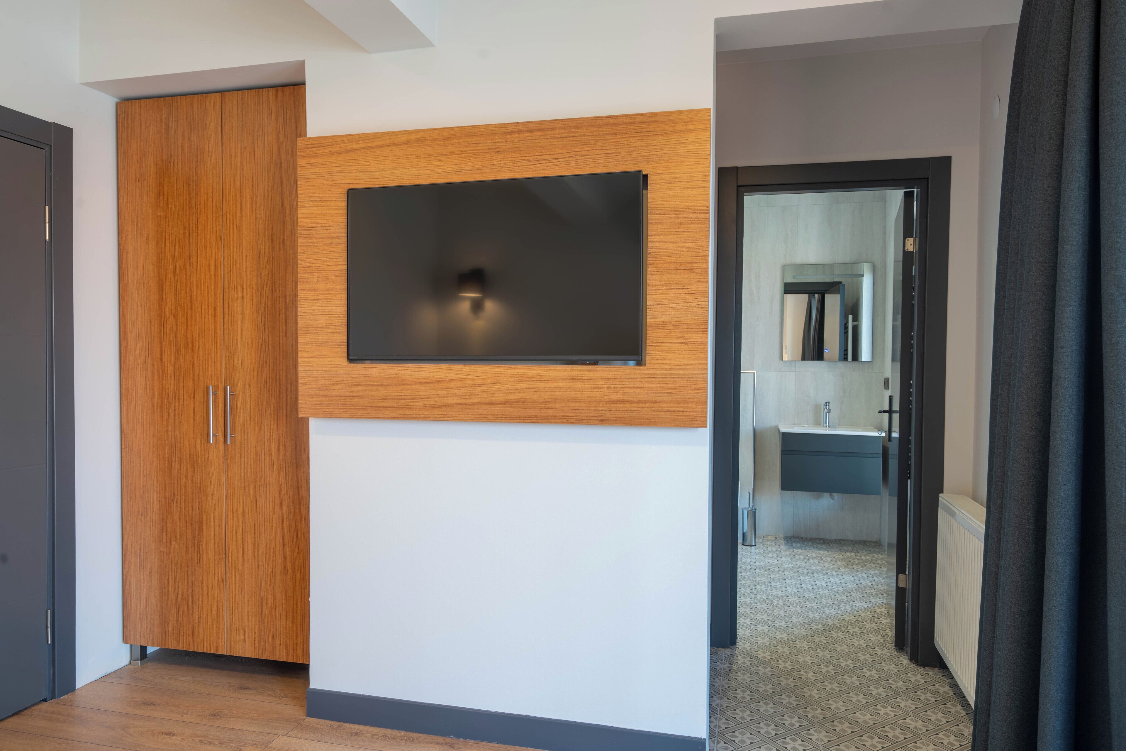 A modern room with a wooden wardrobe, a mounted flat-screen TV, and a bathroom visible through an open door.