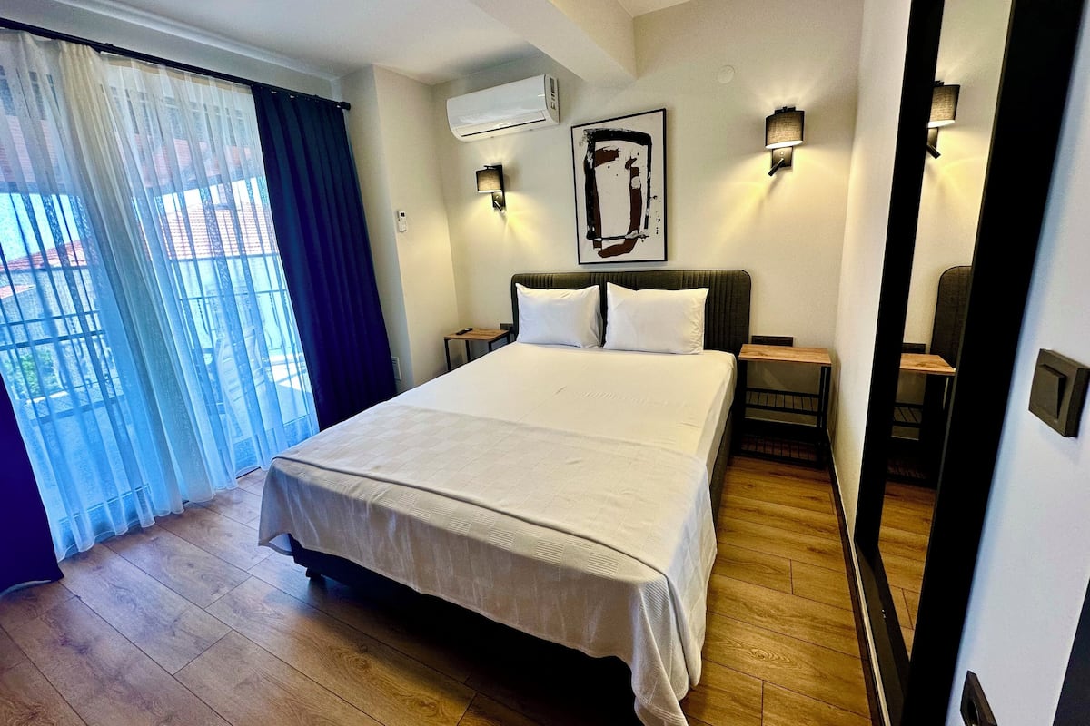Habitación de hotel con una cama grande, una mesita de noche, un equipo de aire acondicionado, una lámpara de pared y una ventana con cortinas.