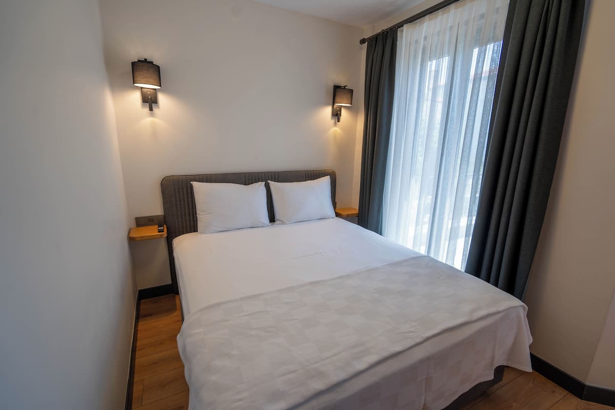 Una cama bien hecha con sábanas blancas, dos lámparas de pared y una ventana con cortinas.