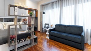 A modern living room with a grey sofa, wooden flooring, a bookshelf with books and decorative items, and a large window with sheer curtains. - The Little Etruscan, cozy tourist rental in the center of Arezzo (Arezzo)