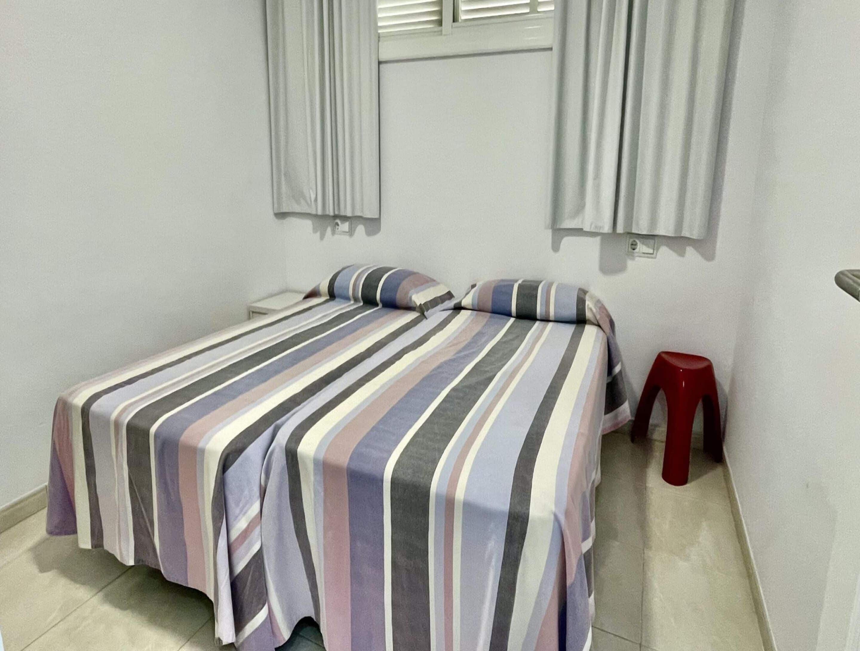 A small, single-bed room with a striped bedspread, a red stool, and a window with blinds.