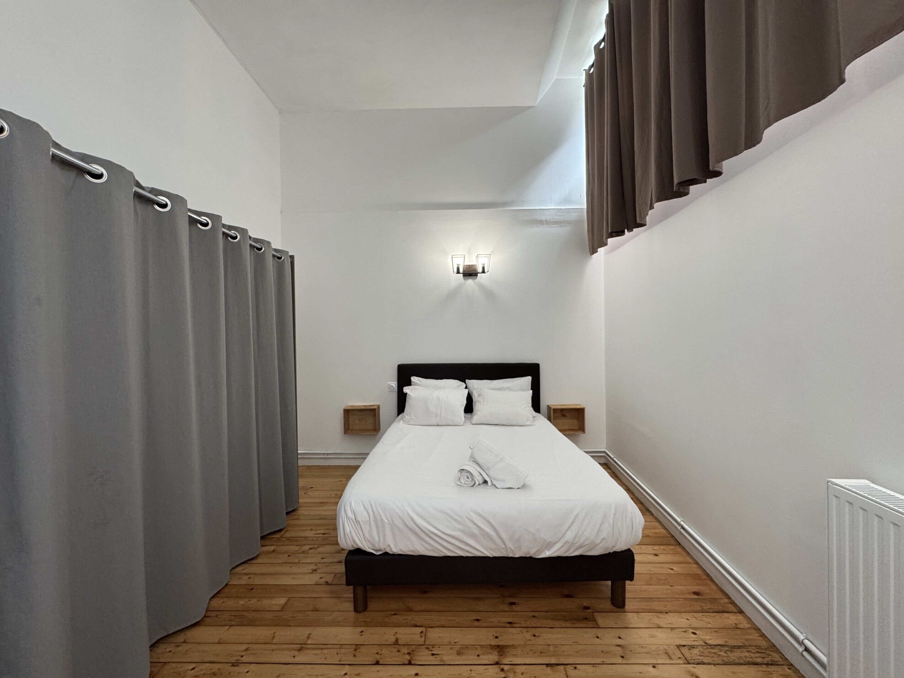 A narrow hotel room with a single bed, wooden flooring, and a small bedside table.