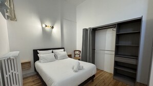 Una cama bien hecha con sábanas blancas, suelo de madera, un radiador, una silla y un armario empotrado.