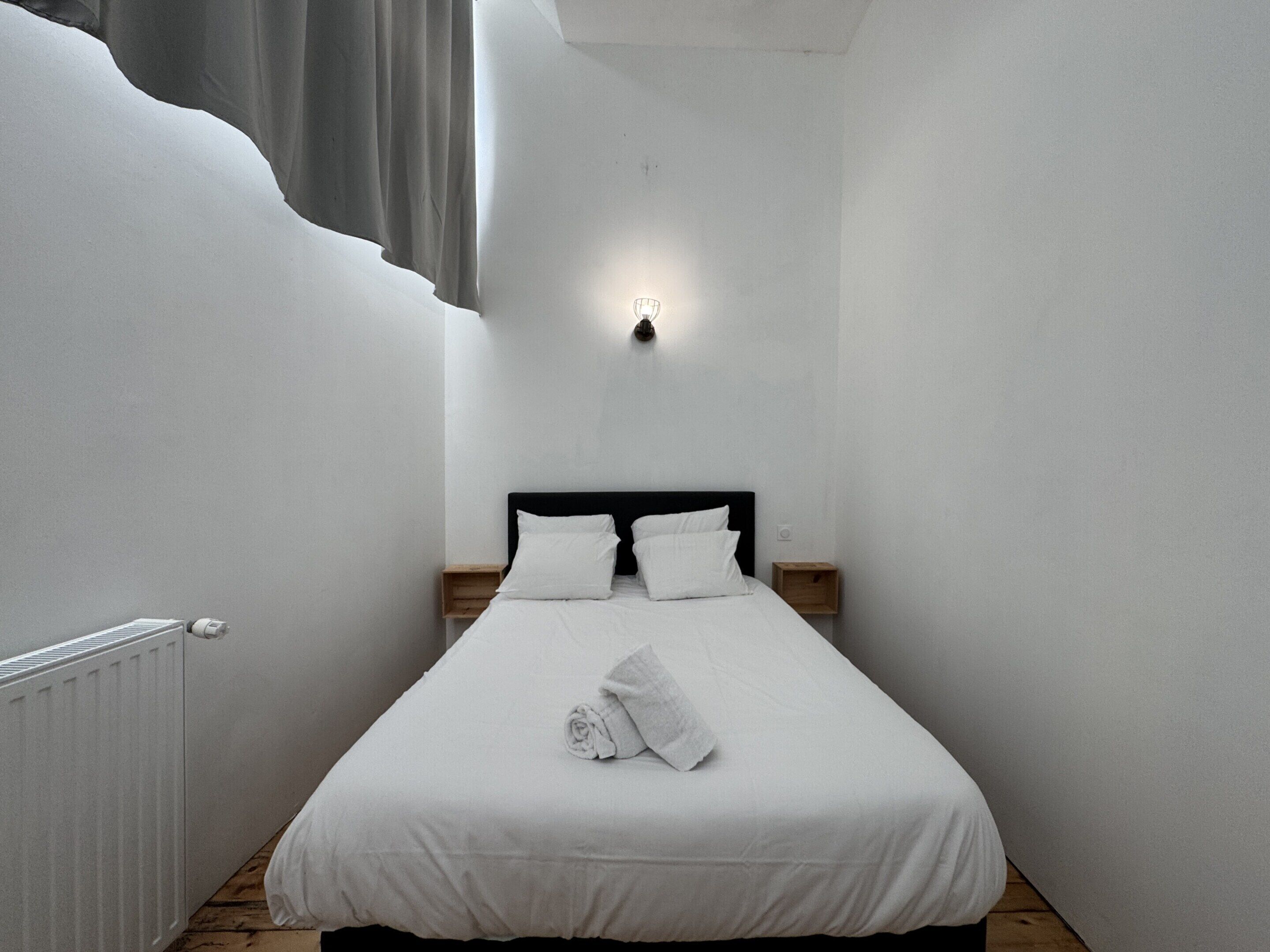 A narrow, white-walled room with a single bed, a nightstand, and a wall-mounted light.