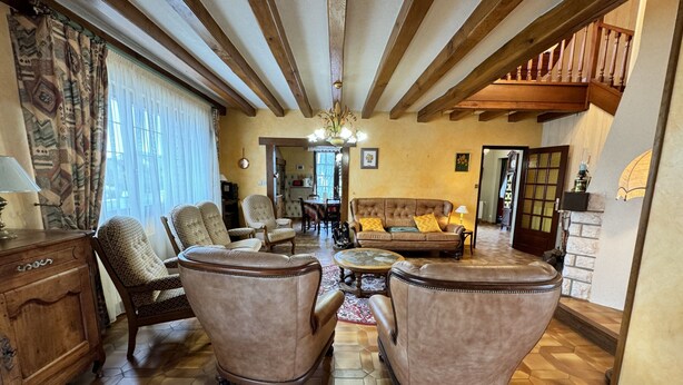 Un salón amplio con techo alto, vigas de madera y chimenea. Hay varios sillones de cuero, un sofá y una mesa de comedor con sillas. El piso es de baldosas y hay un ventanal grande con cortinas.