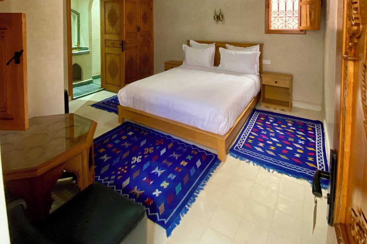 A bedroom with a large bed, wooden headboard, two bedside tables, a nightstand, a decorative ceiling, and a window with bars.
