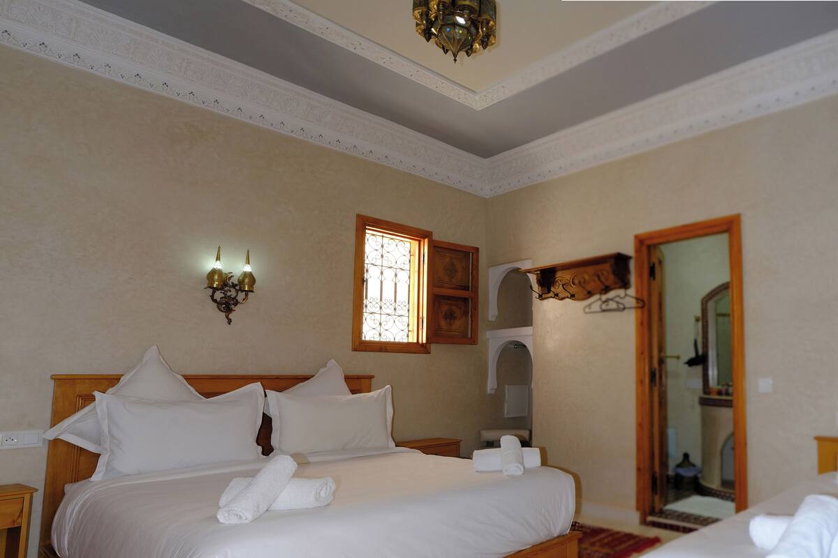 A hotel room with a large bed, a wooden headboard, a small window with decorative iron bars, a wall-mounted light fixture, and a mirror.