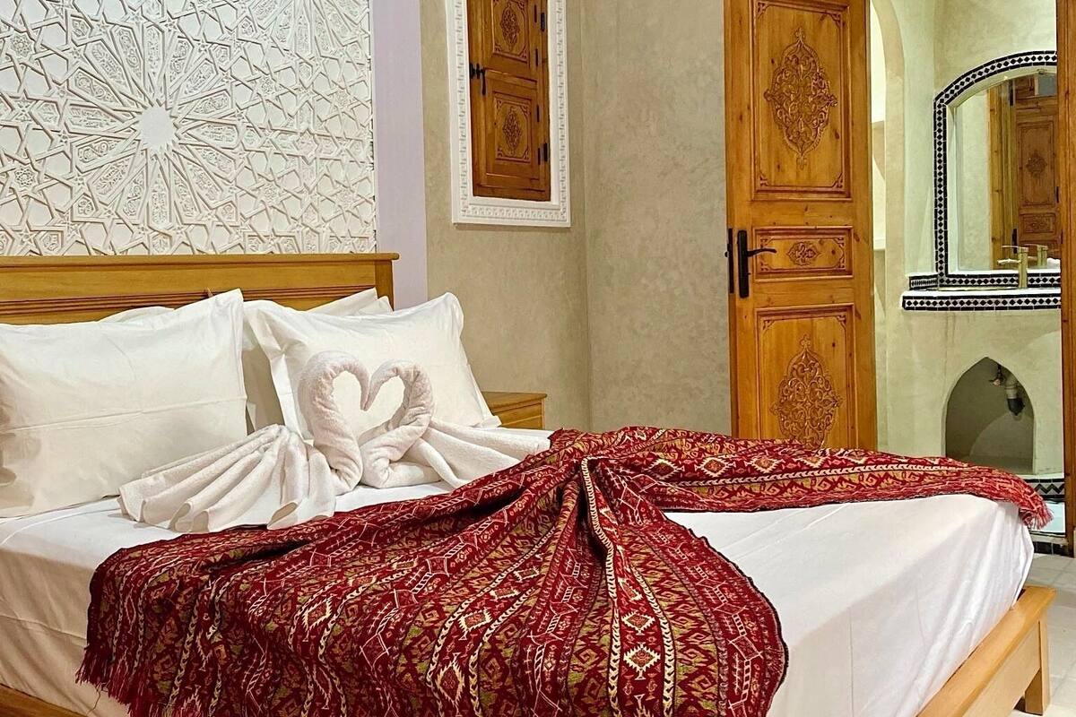 A double bed with white linens and a red patterned throw blanket. There is a wooden door with intricate carvings and a mirror with a black and white border.