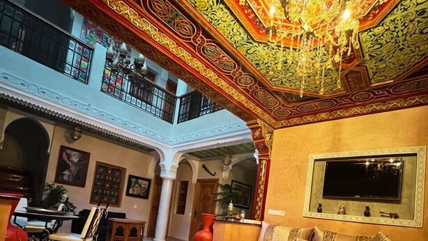 A dining area with a high ceiling and ornate decorations, a television mounted on the wall, and a balcony with a railing. - Riad Rose Mati (Marrakech)