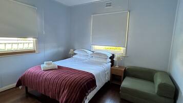 A bedroom with a bed, bedside tables, a nightstand, a green armchair, and a window with blinds.