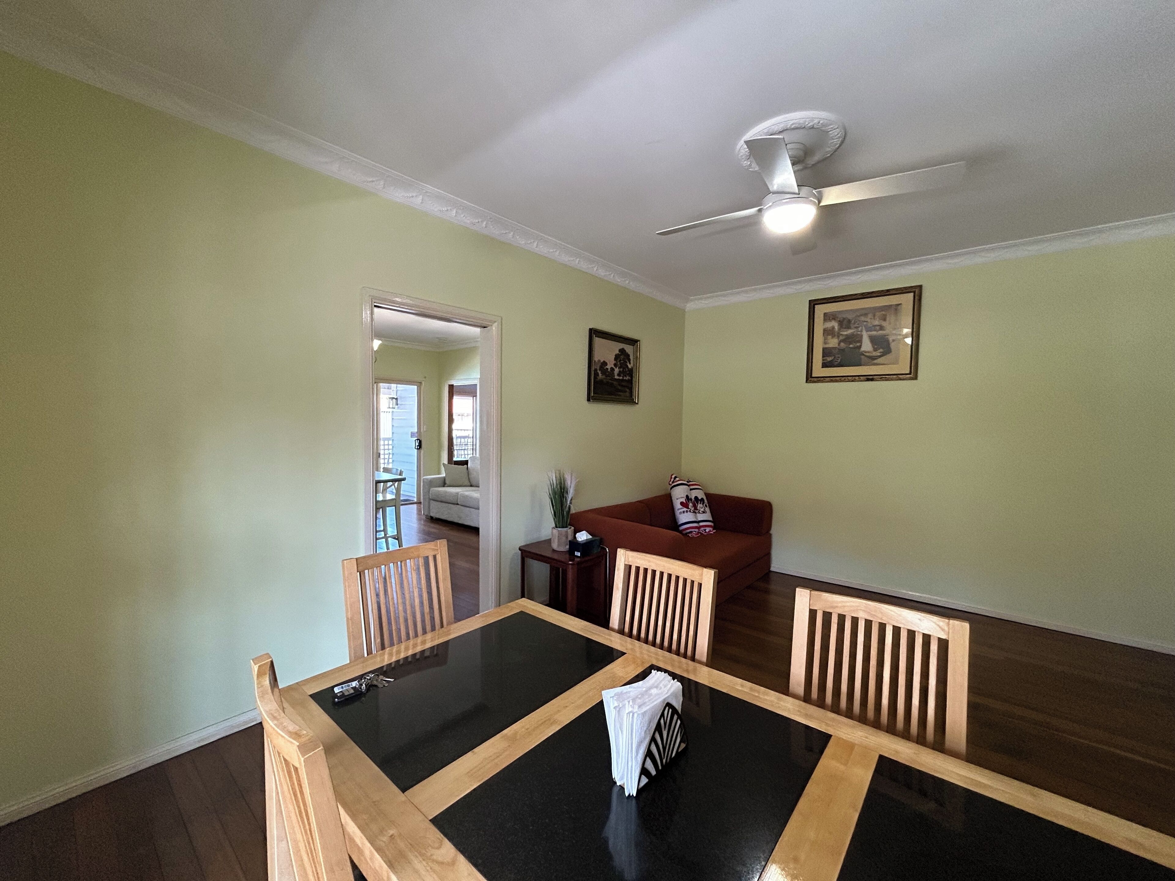 A dining area with a glass-top table, wooden chairs, a ceiling fan, and a view into another room.