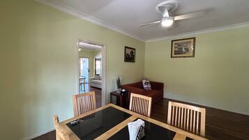 A dining area with a glass-top table, wooden chairs, a ceiling fan, and a view into another room.