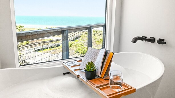 A bathtub with a wooden tray, a book, a glass of water, and a potted plant, with a view of the ocean through a window. - Luxury Ocean Front, Near Downtown, Heated Pool, Hot Tub, Game Room, Sleeps 28 (St. Augustine)