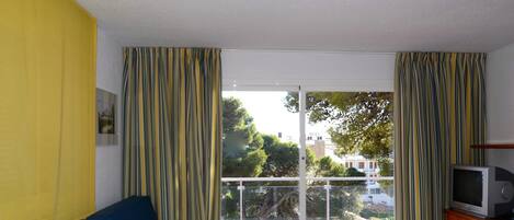 A room with a blue sofa, a wooden table with a television, and a balcony with curtains.