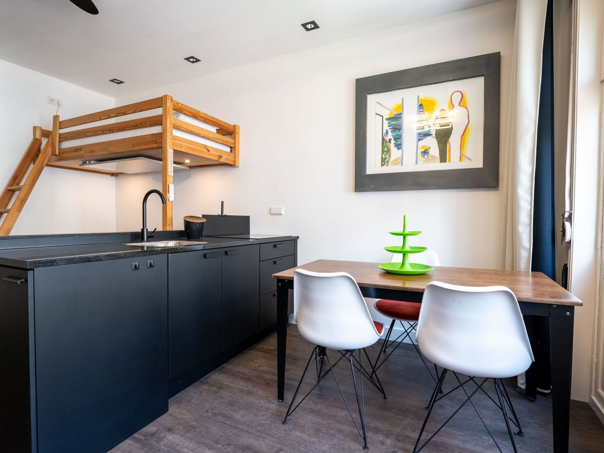 Une cuisine moderne avec des meubles de couleur sombre, une mezzanine en bois et un coin repas agrémenté de deux chaises blanches et d’un centre de table vert.