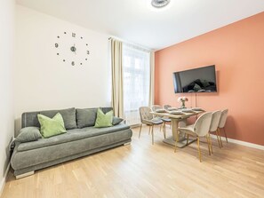 Un salon moderne avec un canapé gris, une table à manger avec des chaises, un téléviseur à écran plat fixé au mur et une grande fenêtre avec des rideaux.