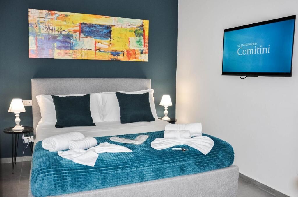 A hotel room with a bed, pillows, a blue blanket, a television, and a painting on the wall.