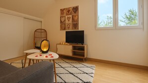 A living room with a sofa, a small round table with rose petals, a television on a wooden cabinet, and a window with a view of trees. - Cosy nest in SAINT VALERY EN CAUX near the sea. (Saint-Valery-en-Caux)