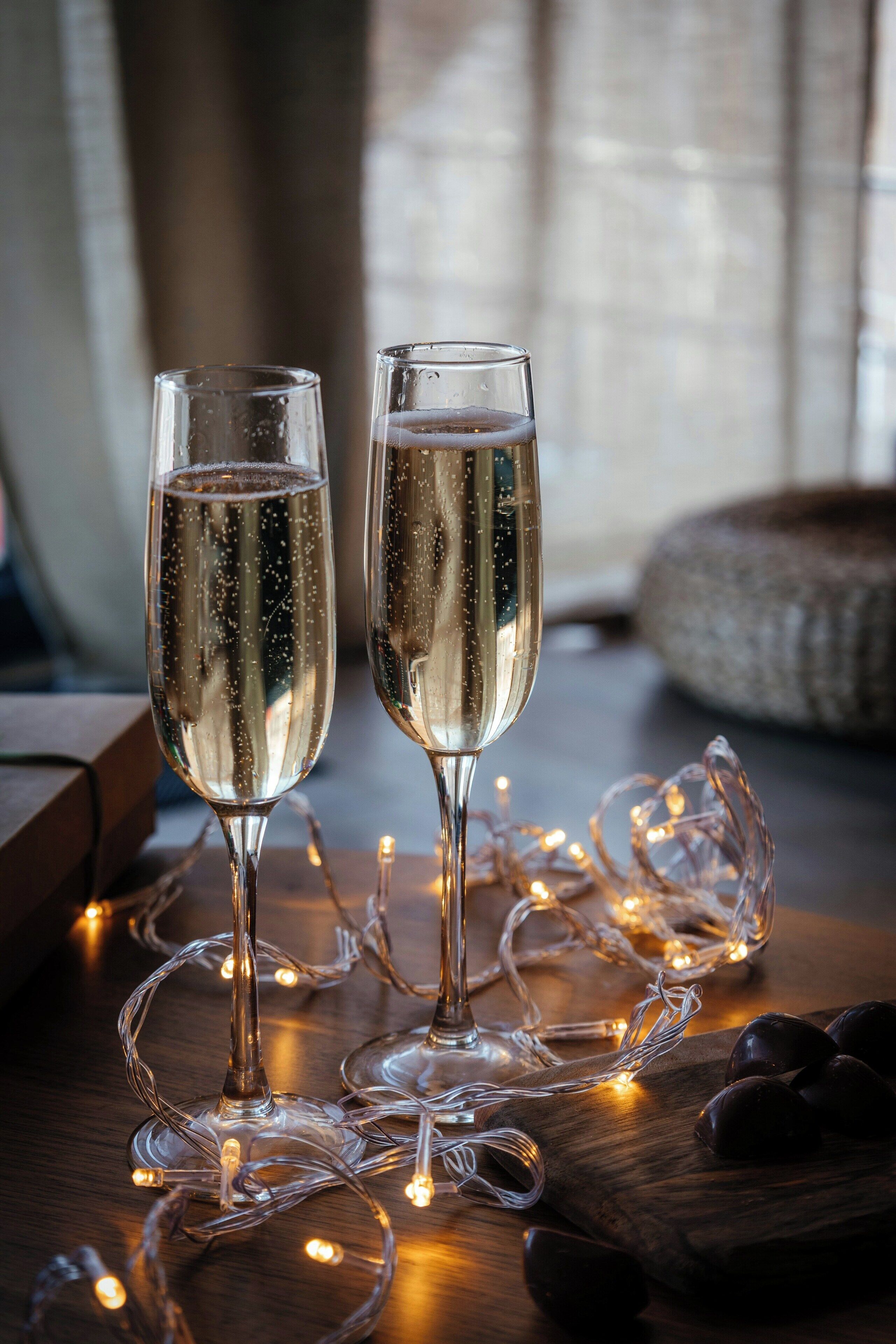 Two champagne flutes on a wooden surface with string lights and dark round objects, possibly chocolates, in a dimly lit setting.