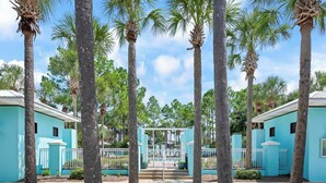 A gated entrance with palm trees, a blue building, and a clear sky. - Gulf Place 30A! Walk to Beach! Pools! Tennis Court! (Santa Rosa Beach)