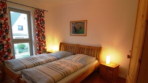 A double bed with a wooden headboard, two bedside tables with lamps, a window with floral curtains, and a framed picture on the wall. - Hamburg Wedel Apartment NORDLICHT on the Elbe (Wedel)