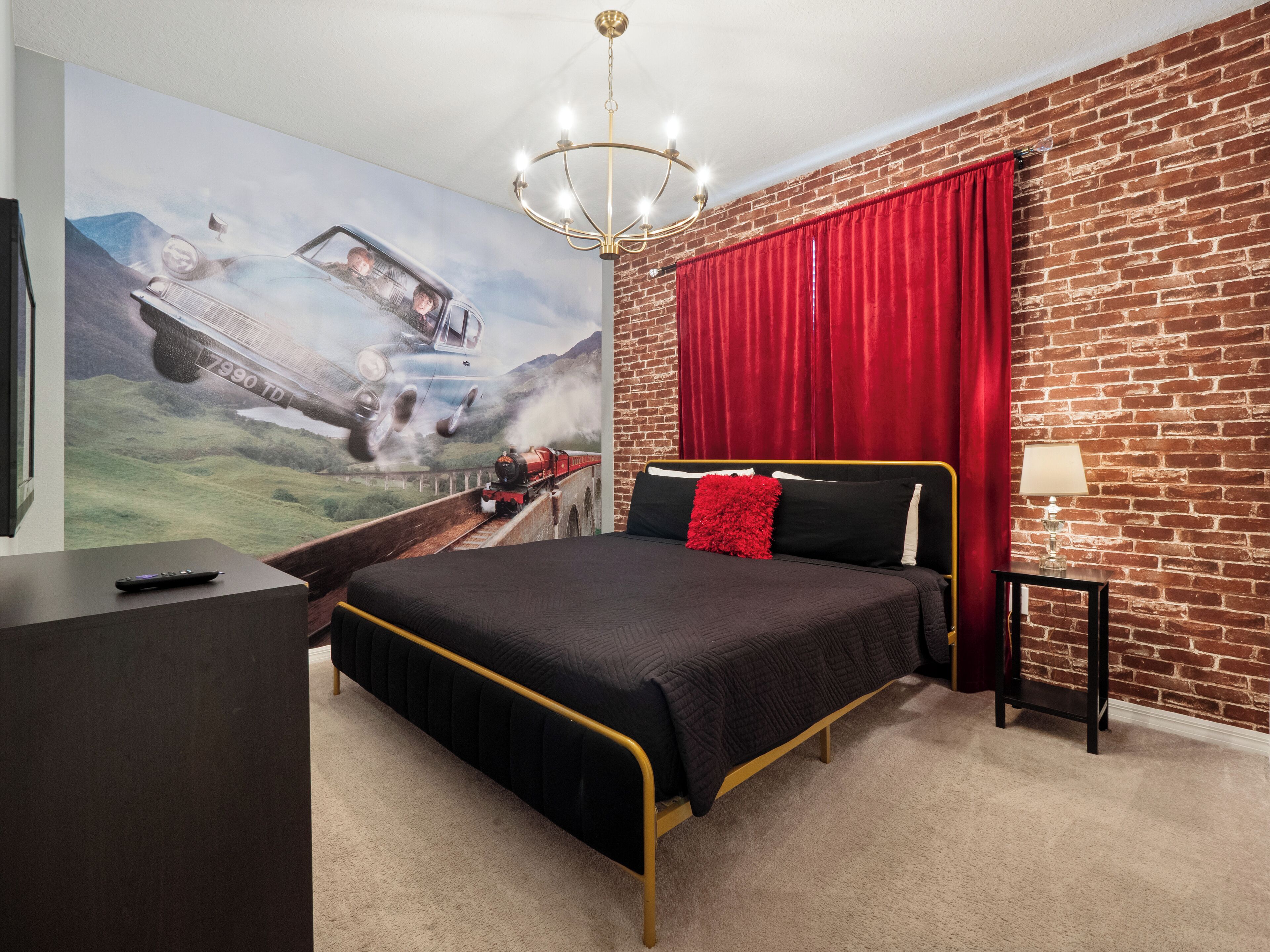 A bedroom with a brick wall, a large window with red curtains, a bed with black bedding and a red pillow, a chandelier, and a TV on a stand.