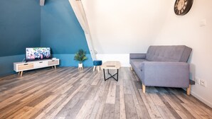 A modern living room with a sloped ceiling, a grey sofa, a wooden TV stand, a small table, and a potted plant. - Chic Apartment Near City Center (Ecommoy)