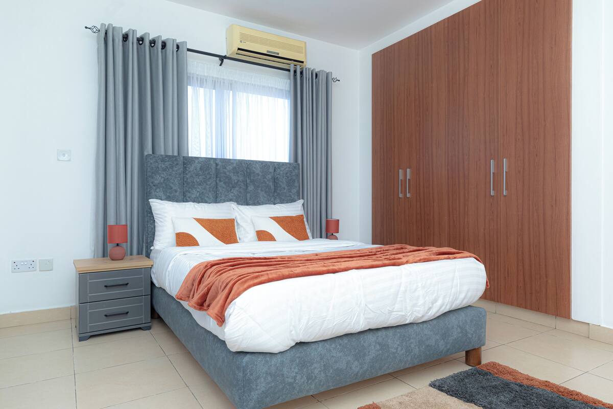 A modern bedroom with a bed, grey curtains, a wooden wardrobe, and a nightstand with a lamp.