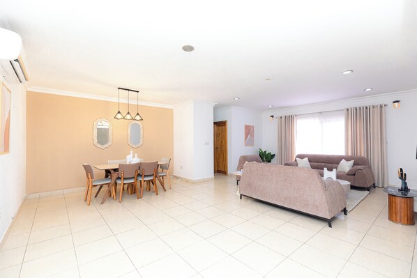 A modern living room with a beige sofa, a dining table with chairs, and a large window with curtains. - Polo Heights -Sky Suites (Accra)