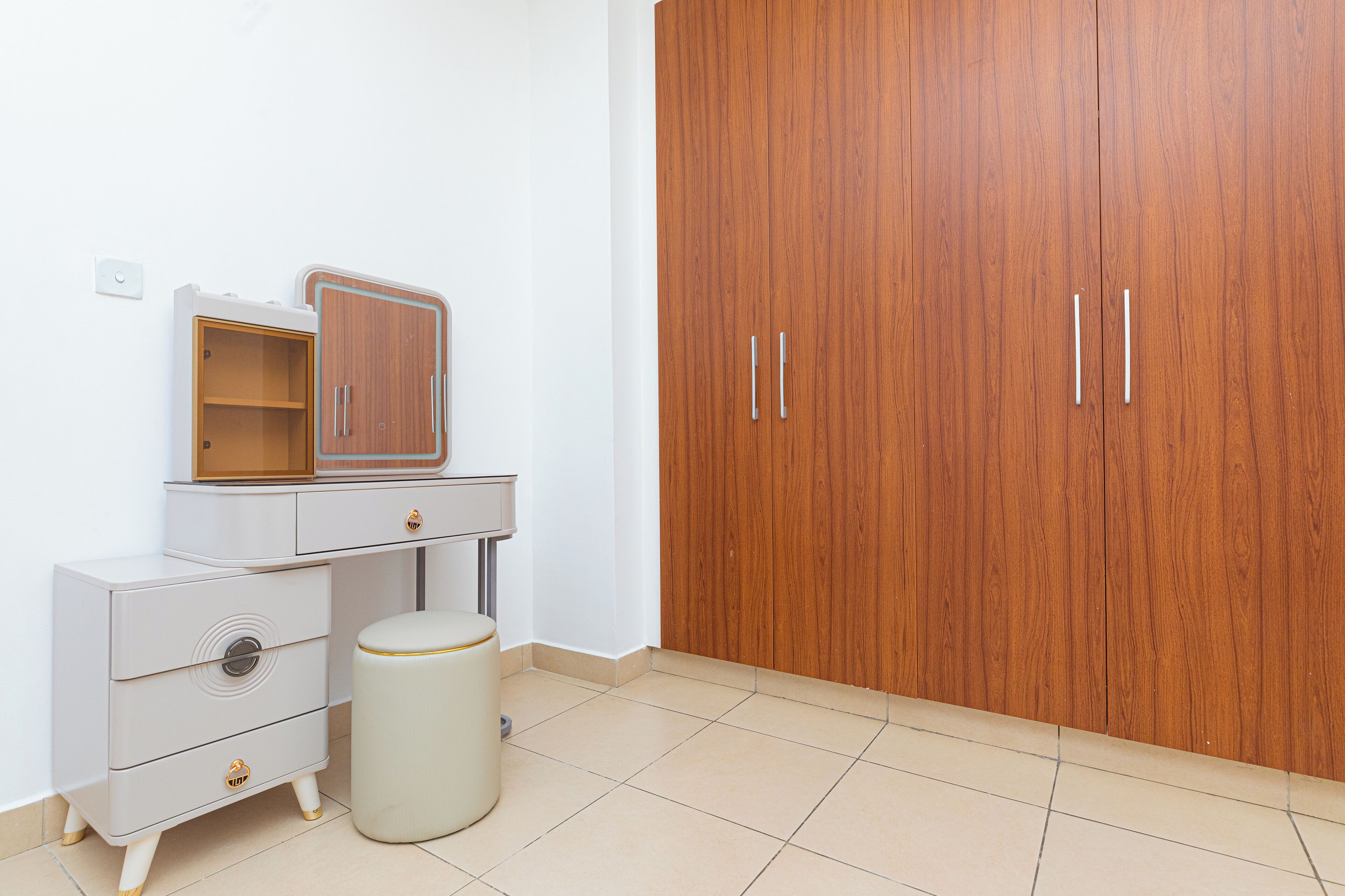 Una habitación con un armario de madera, un tocador blanco con espejo y un taburete pequeño y redondo.