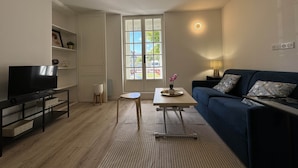 A modern living room with a flat-screen TV, a blue sofa, a small table, and a large window with a view of greenery. - Apartment in Le Mans With Private Terrace (Le Mans)