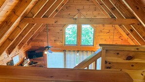 A wooden attic room with a sloped ceiling, a ceiling fan, and a window overlooking greenery.