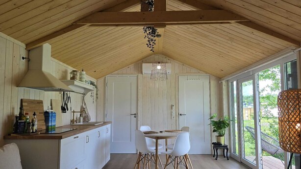 A cozy kitchen with wooden beams, a dining area with white chairs, and large windows offering a view of the outdoors. - Tiny House Drents-Friese Wold with terrace and parking lot (Hoogersmilde)