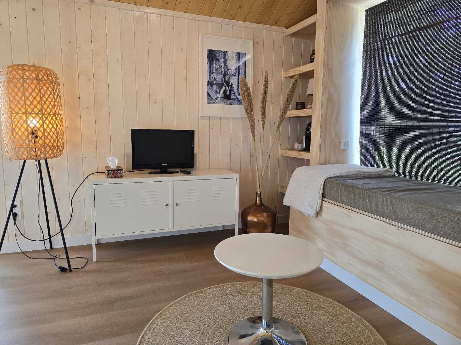 Een knus interieur met een houten vloer, een witte kast met een televisie, een ronde tafel en een bed met een raam.